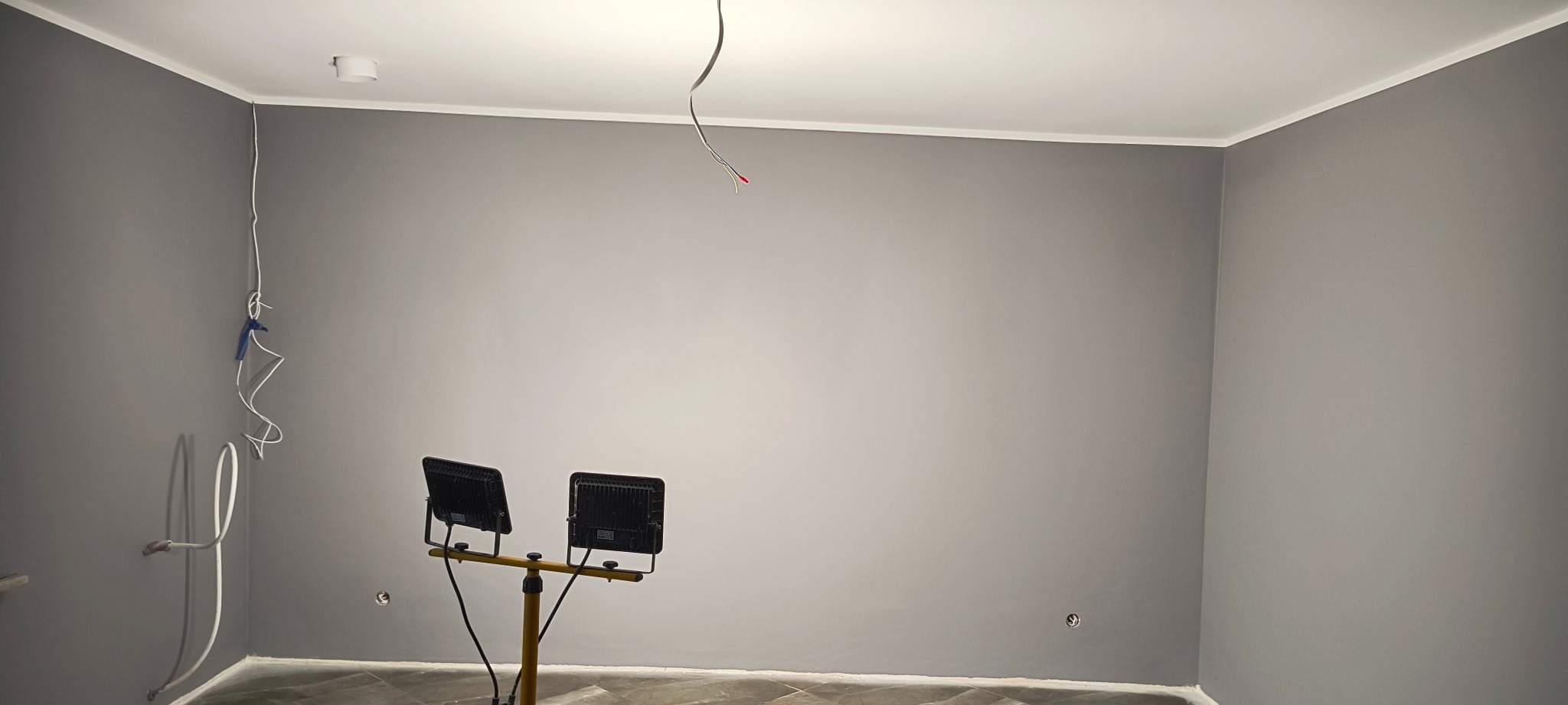 Pokój po malowaniu na szaro, widoczne przewody elektryczne, lampy halogenowe na statywie i fragment podłogi