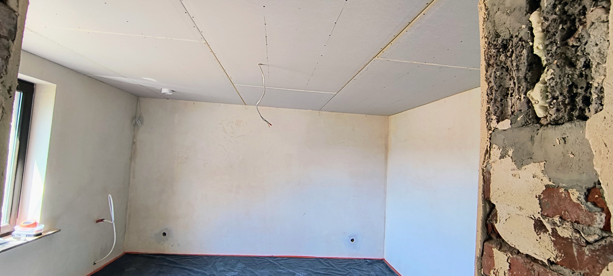 Pokój w trakcie prac wykończeniowych: sufit podwieszany z płyt kartonowo-gipsowych, ściany po wstępnym wyrównaniu, widoczne instalacje elektryczne i hydrauliczne, zabezpieczona podłoga, fragment...