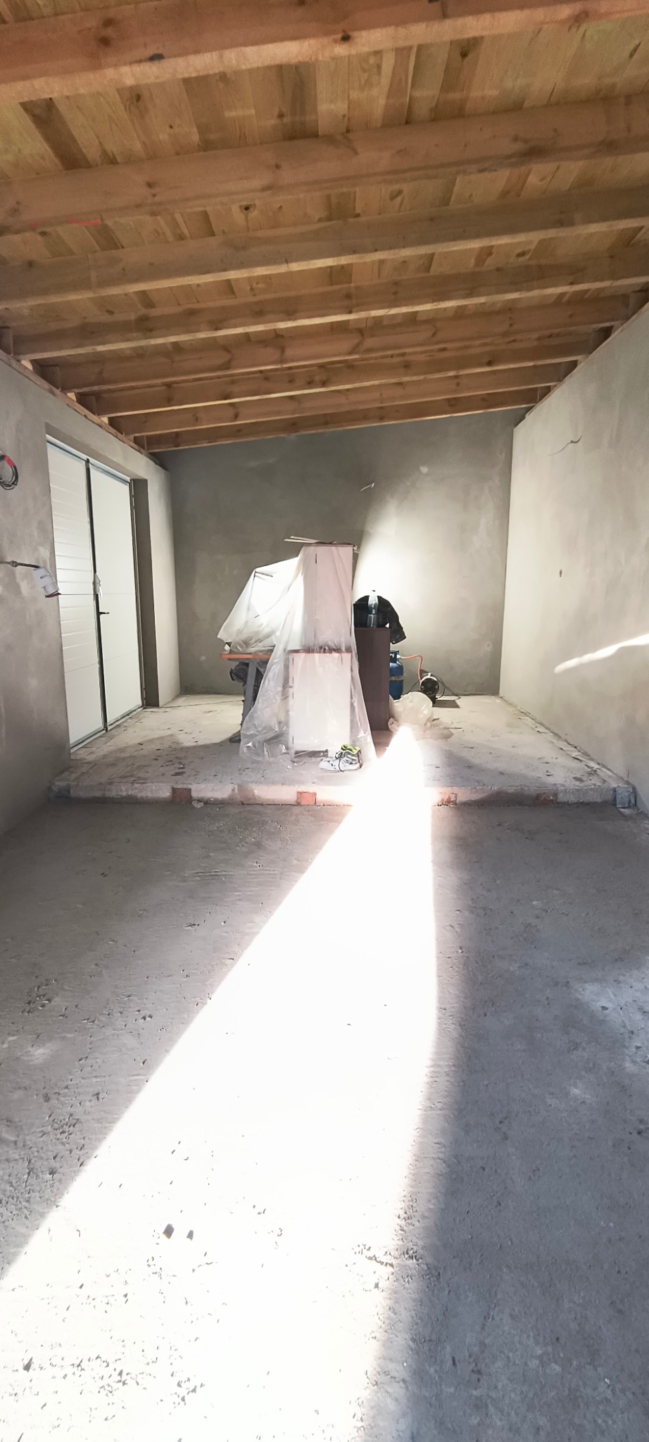 Wnętrze pomieszczenia w trakcie prac wykończeniowych, widoczne surowe ściany, drewniany strop, zabezpieczone meble folią i sprzęt budowlany.