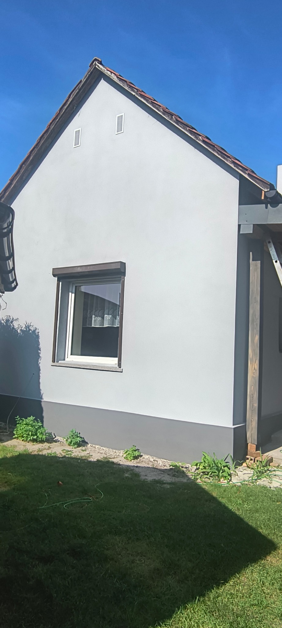 Otynkowana elewacja domu jednorodzinnego w odcieniach szarości, z widocznym fragmentem dachu z dachówką, oknem z roletą i elementami ogrodu.