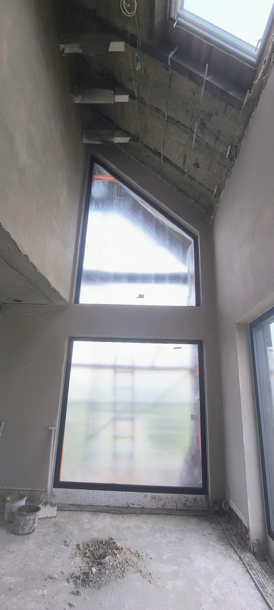 Wnętrze w trakcie prac wykończeniowych, ściany pokryte świeżym tynkiem, okna zabezpieczone folią, widoczne niedoskonałości i pozostałości zaprawy na podłodze, nietypowy kształt okna dachowego.