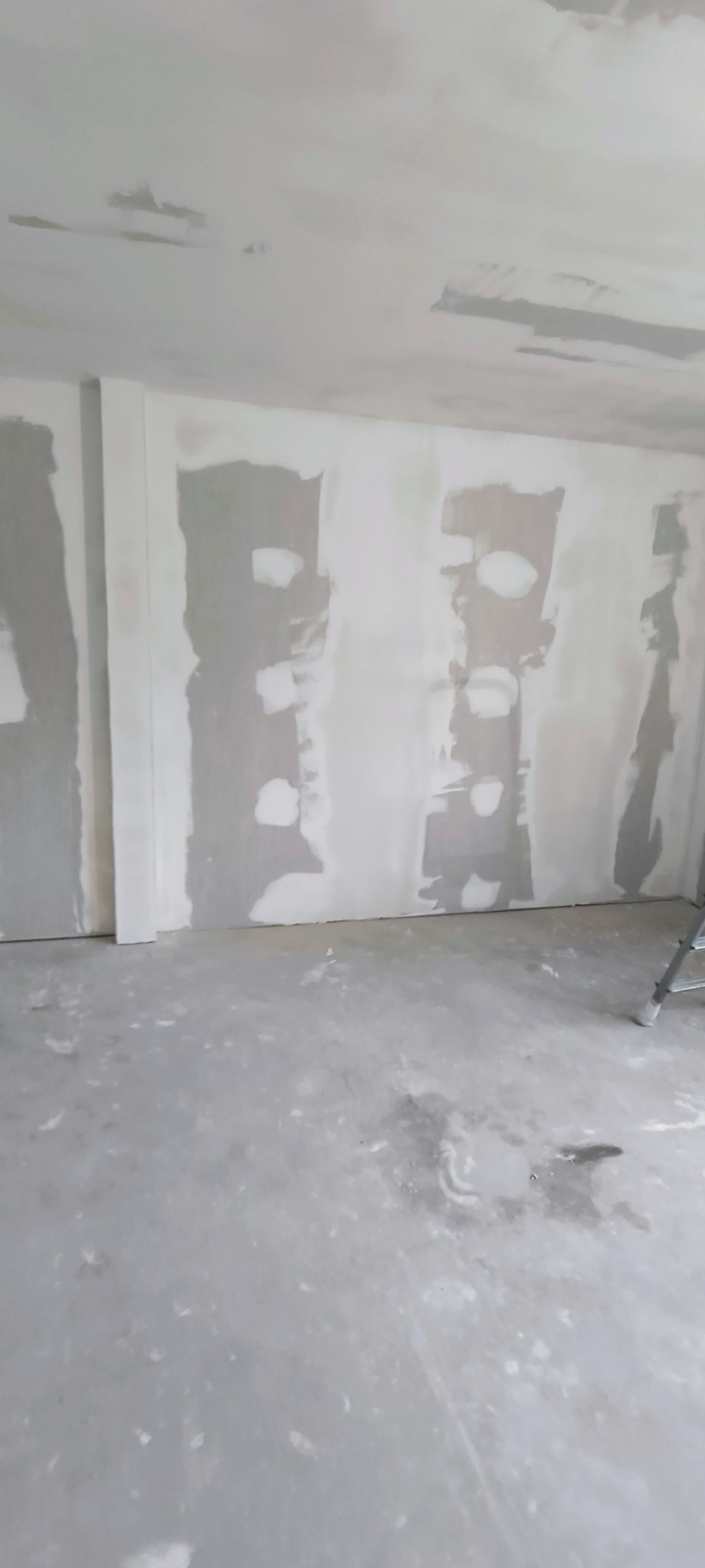 Wnętrze pokoju w trakcie prac wykończeniowych, ściany częściowo pokryte szpachlą, widoczne łączenia płyt gipsowo-kartonowych, podłoga zabezpieczona przed zabrudzeniem, fragment drabiny w rogu.