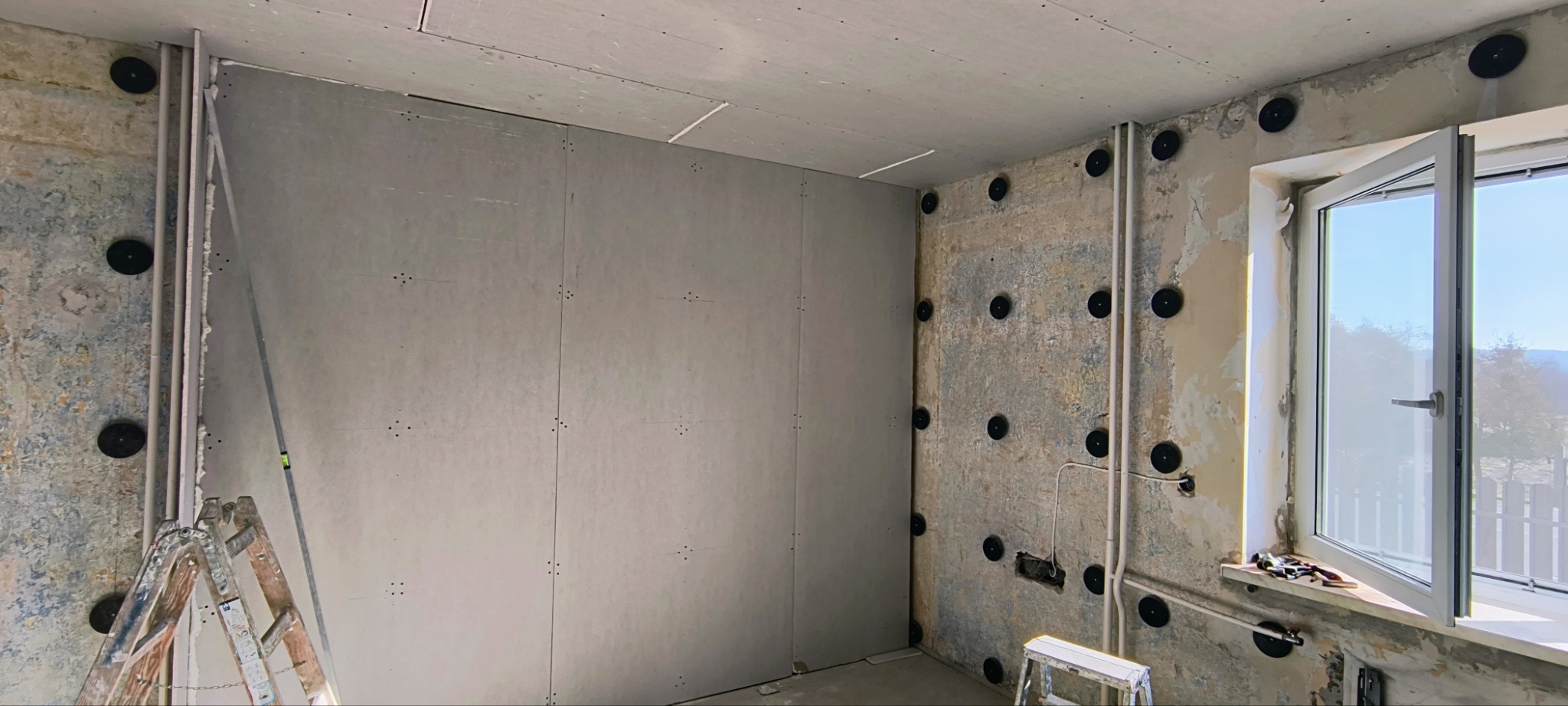 Wnętrze w trakcie prac wykończeniowych: ściany z płyt gipsowo-kartonowych, widoczne mocowania, fragment drabiny, okno z widokiem na zewnątrz.