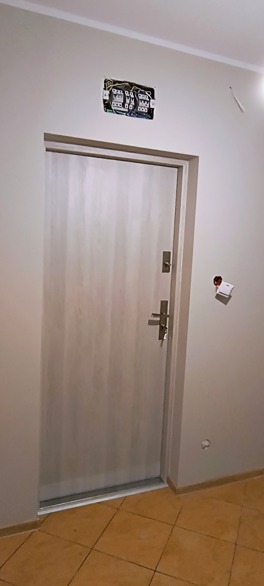 Drzwi wejściowe w trakcie remontu, widoczna skrzynka elektryczna bez osłony i wystające przewody.