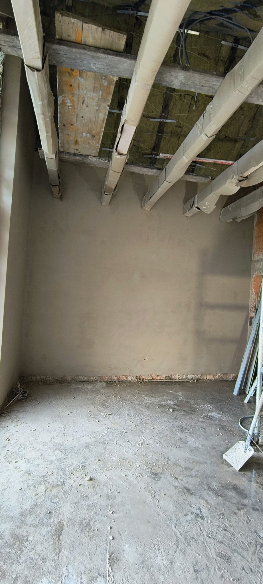 Świeżo nałożony tynk maszynowy na ścianie w pomieszczeniu w trakcie remontu, widoczne zabezpieczone elementy konstrukcyjne sufitu i resztki materiałów budowlanych na podłodze.