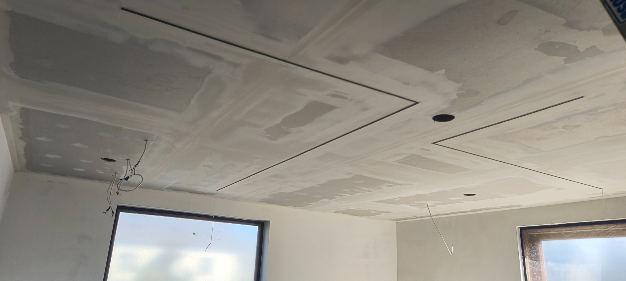 Sufit z płyt gipsowo-kartonowych po szpachlowaniu, widoczne łączenia i otwory na oświetlenie, czarne listwy dekoracyjne, okna z widokiem na zewnątrz.