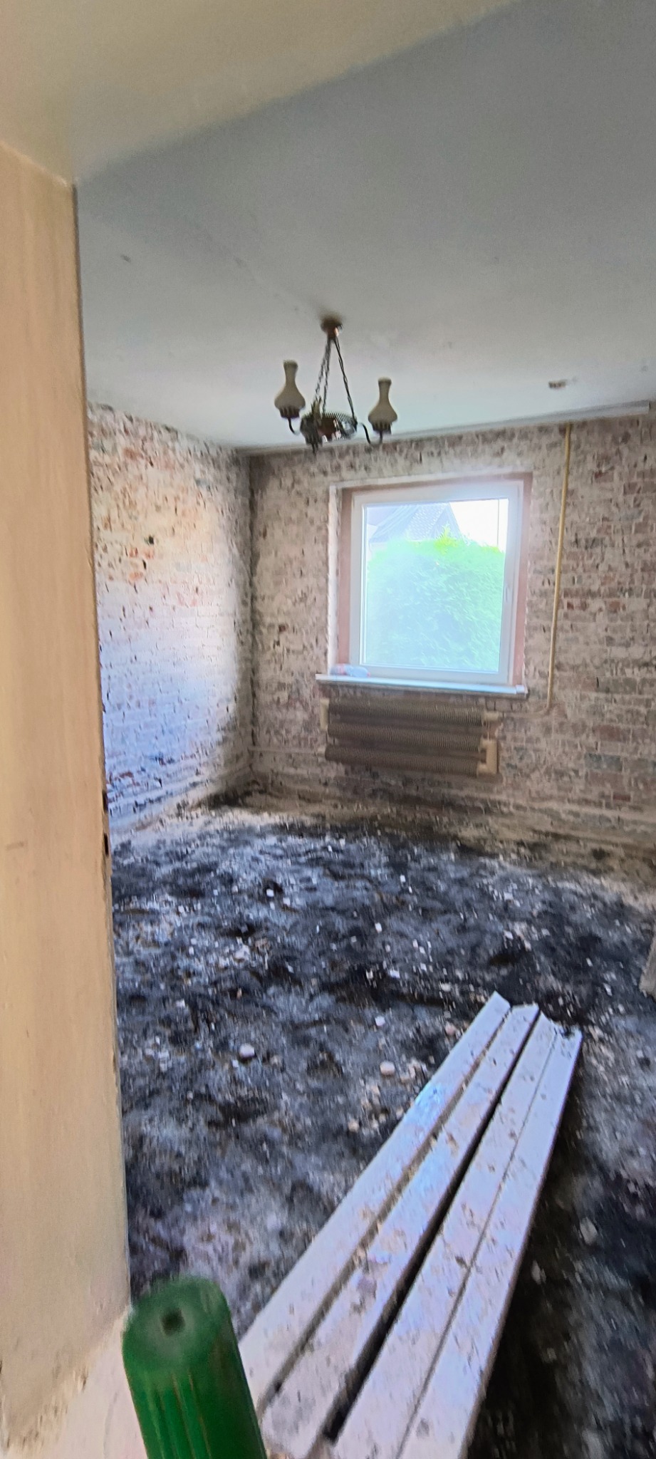 Pokój w trakcie remontu: surowe cegły na ścianach, okno z widokiem na zieleń, stara lampa wisząca i zabrudzone podłoże z leżącymi listwami przypodłogowymi.
