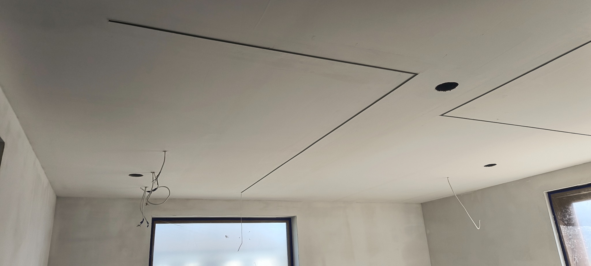 Świeżo położona gładź na suficie z wpuszczonymi liniami oświetleniowymi i otworami na punkty świetlne, widoczne kable i surowe ściany.