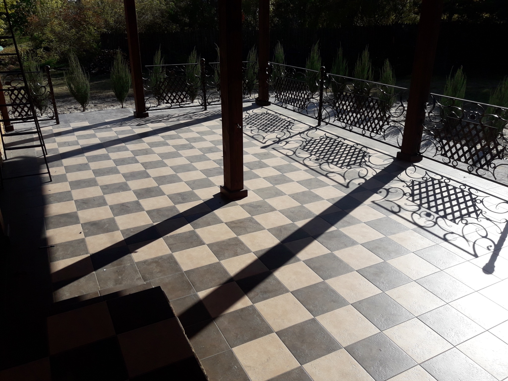 Szachownica z płytek na zadaszonym tarasie, rzucająca cień z drewnianych słupów i ozdobnej, metalowej balustrady. Widok na zieleń w tle.