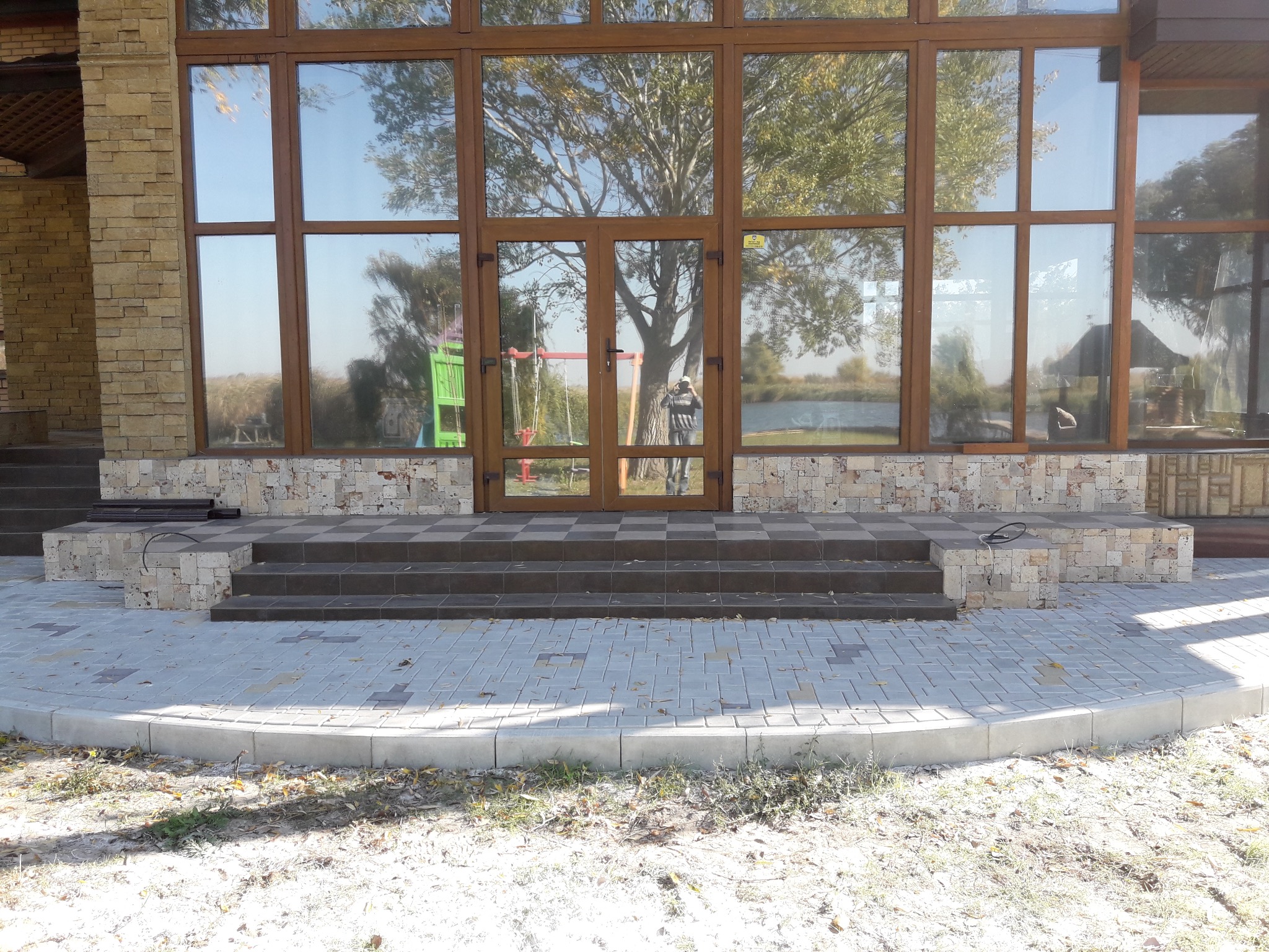 Wykończony taras z kostki brukowej i schodami w Tereni. Duże okna z drewnianą ramą odbijają drzewa i jezioro. Kamienna elewacja budynku.