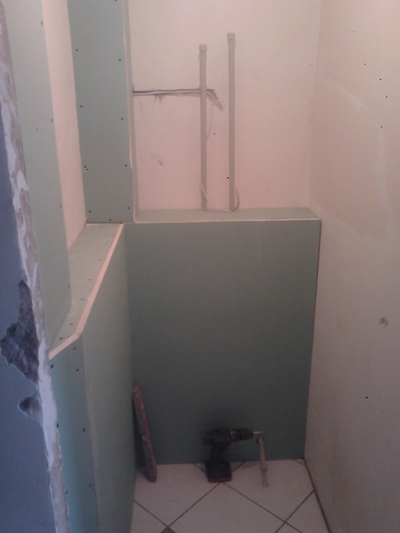 Montaż płyt gipsowo-kartonowych w małej łazience, widoczne profile i narzędzia. Częściowo wykończona ściana z instalacją hydrauliczną w tle.