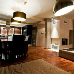 INVOST SPÓŁKA Z OGRANICZONĄ ODPOWIEDZIALNOŚCIĄ - Przestronny salon z drewnianą podłogą, kominkiem obłożonym beżową płytką imitującą kamień, stołem z krzesłami oraz kuchnią w tle, oświetlony lampami wiszącymi.