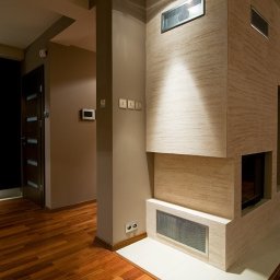 INVOST SPÓŁKA Z OGRANICZONĄ ODPOWIEDZIALNOŚCIĄ - Nowoczesne wnętrze z drewnianą podłogą, kolumną i kominkiem wykończonym beżowym tynkiem strukturalnym, oświetlonym punktowym światłem z sufitu.