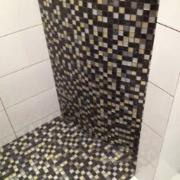 Wykończona wnęka prysznicowa z mozaiką w odcieniach czerni, szarości i złota, kontrastująca z białymi, prostokątnymi płytkami na ścianach.