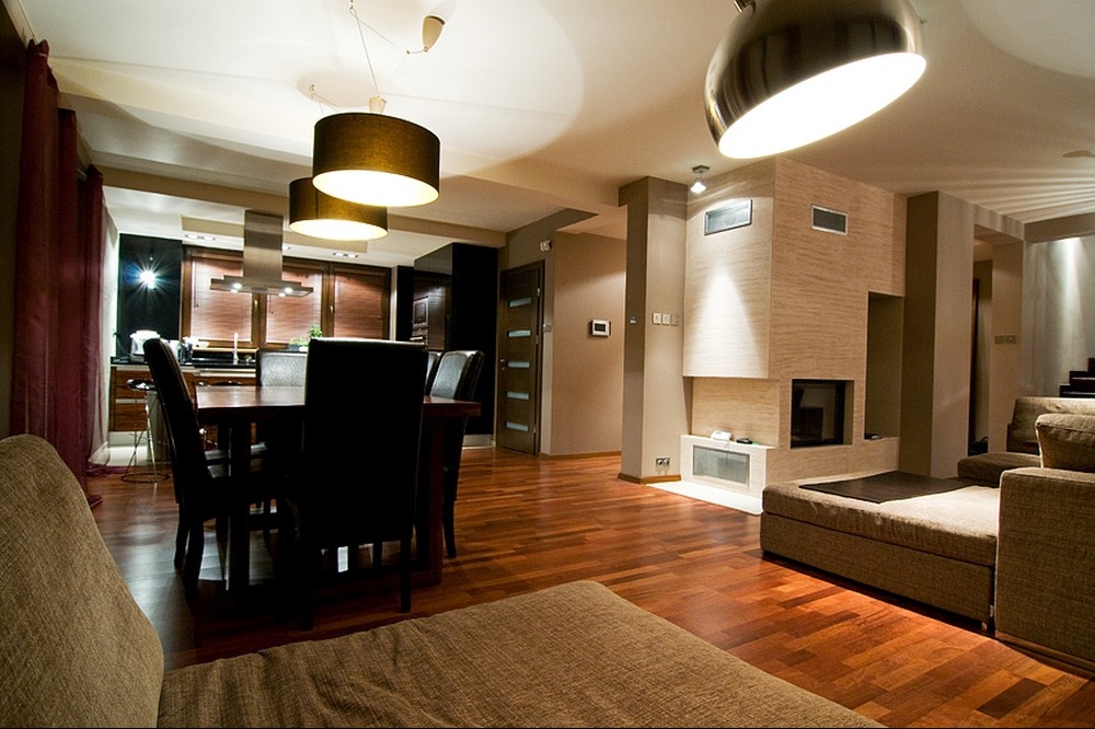 Przestronny salon z drewnianą podłogą, kominkiem obłożonym beżową płytką imitującą kamień, stołem z krzesłami oraz kuchnią w tle, oświetlony lampami wiszącymi.