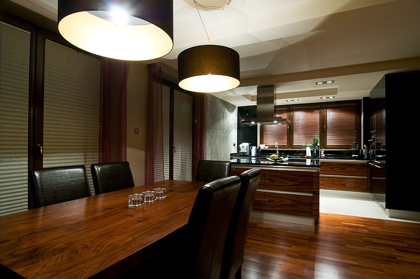 Nowoczesna jadalnia z drewnianym stołem, skórzanymi krzesłami i otwartą kuchnią w tle, oświetlona wiszącymi lampami z abażurami.