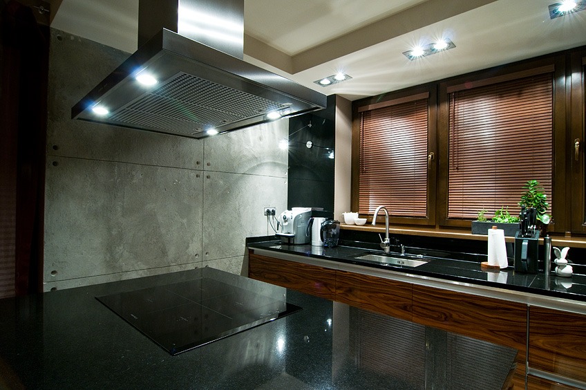 Nowoczesna kuchnia z betonową ścianą, wyspą z czarnym blatem i drewnianymi szafkami, okno z żaluzjami i oświetleniem punktowym.