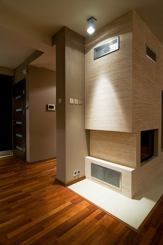 Nowoczesne wnętrze z drewnianą podłogą, kolumną i kominkiem wykończonym beżowym tynkiem strukturalnym, oświetlonym punktowym światłem z sufitu.