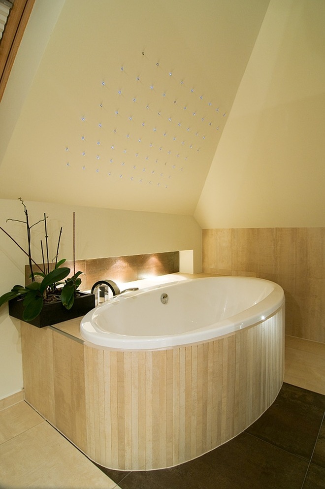 Owalna wanna obudowana panelami imitującymi drewno w łazience na poddaszu w Katowicach, oświetlona punktowymi lampami w suficie, z dekoracyjną rośliną w doniczce.