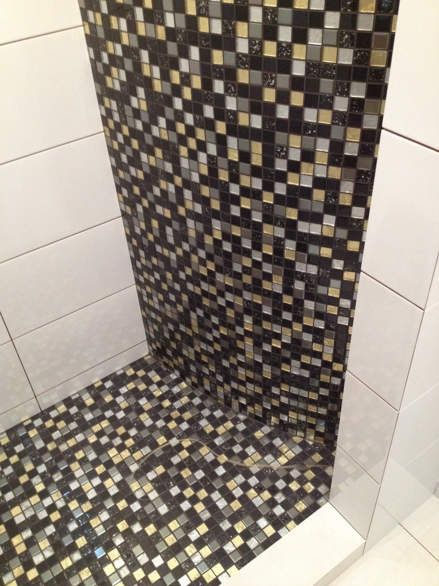 Wykończona wnęka prysznicowa z mozaiką w odcieniach czerni, szarości i złota, kontrastująca z białymi, prostokątnymi płytkami na ścianach.