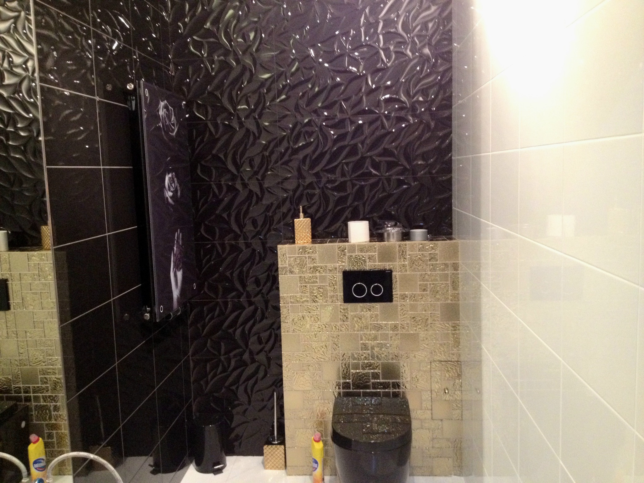 Nowoczesna łazienka z czarną miską WC, mozaiką w odcieniach złota i czarnymi płytkami 3D na ścianie.