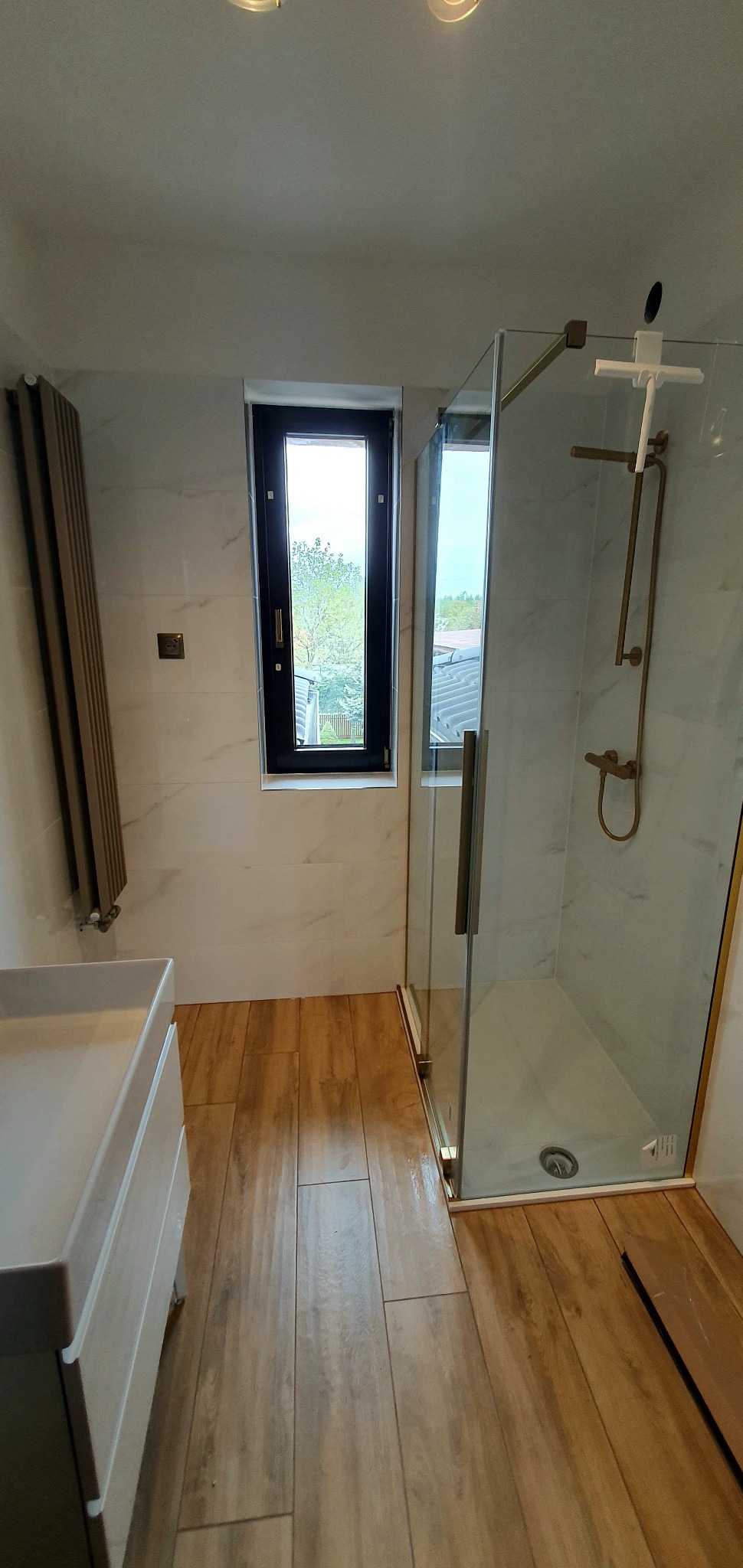 Nowoczesna łazienka z kabiną prysznicową ze złotymi okuciami, umywalką z szafką, grzejnikiem żeberkowym i oknem z czarną ramą.