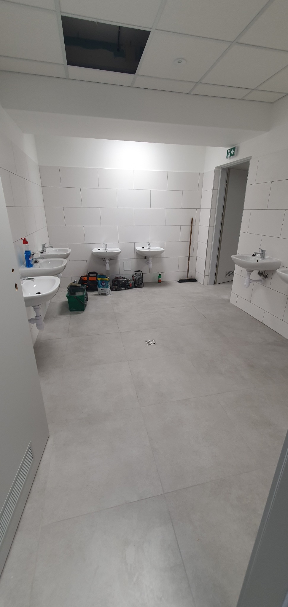 Wnętrze publicznej toalety w trakcie prac wykończeniowych, widoczne umywalki, białe płytki na ścianach, narzędzia i materiały budowlane rozłożone na podłodze, otwarty sufit podwieszany.