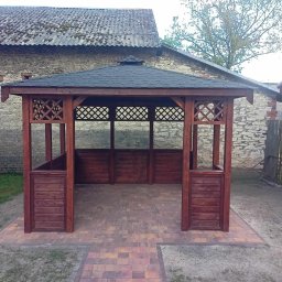 ALBERO Mateusz Kosiń - Drewniana konstrukcja z czarnym dachem, prawdopodobnie altana, na tle kamiennego budynku i kostki brukowej