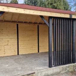 Drewniana wiata z czerwonym dachem, czarnymi słupami i ażurowymi panelami. Konstrukcja z jasnego drewna, betonowa podstawa. Widok na ogród i drzewa.