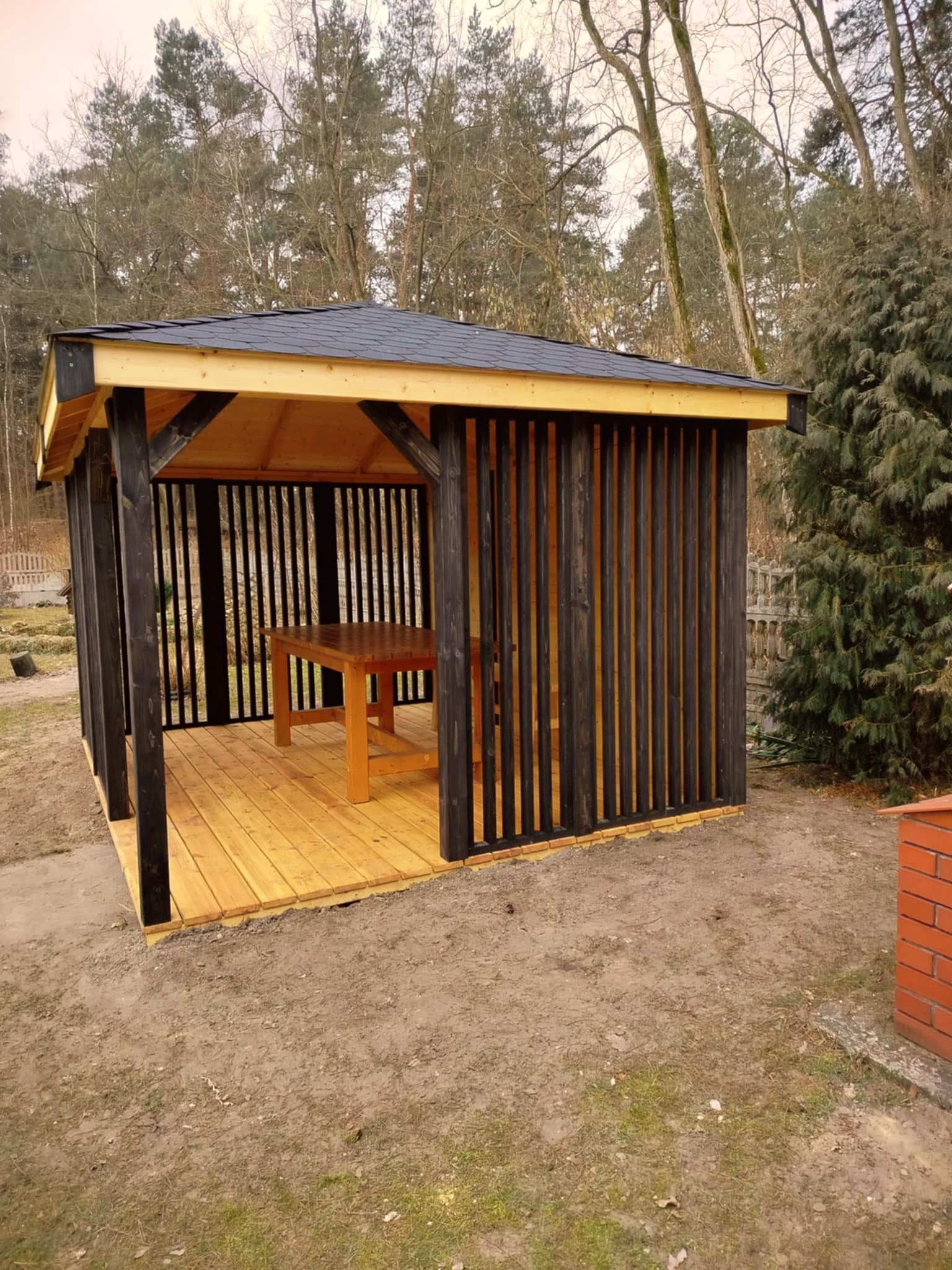 Nowoczesna altana ogrodowa z drewnianą podłogą, stołem i czarnymi pionowymi panelami, na tle drzew bezlistnych i iglaka.