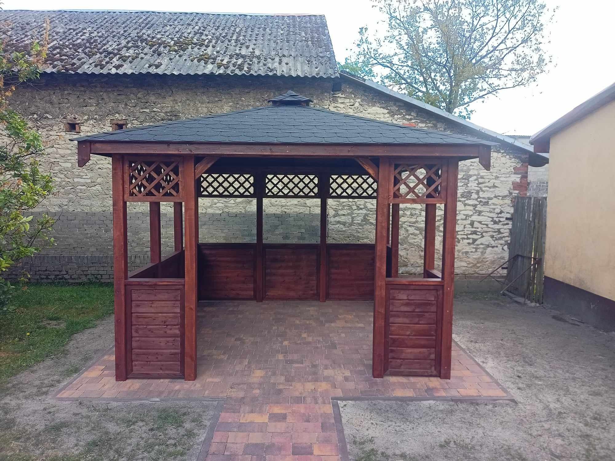 Drewniana konstrukcja z czarnym dachem, prawdopodobnie altana, na tle kamiennego budynku i kostki brukowej