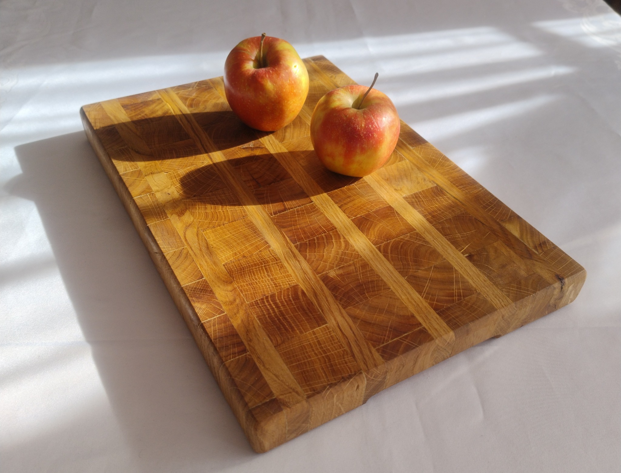 Drewniana deska do krojenia z klejonego drewna dębowego, wykończona olejem, z dwoma czerwono-żółtymi jabłkami leżącymi na powierzchni, na białym tle.