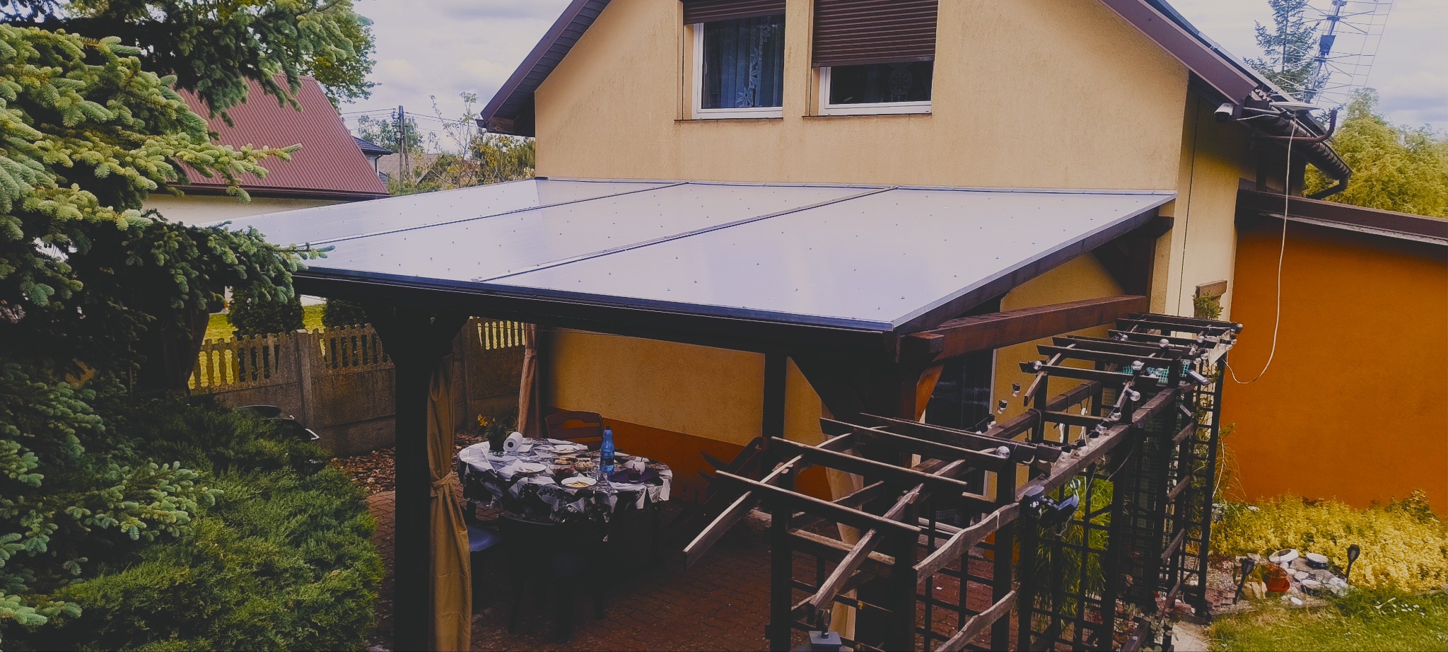 Zadaszona, ciemnobrązowa konstrukcja drewniana przylegająca do budynku, z przezroczystym dachem. Widoczny stół z nakryciem i krzesła. Na pierwszym planie fragment pergoli.