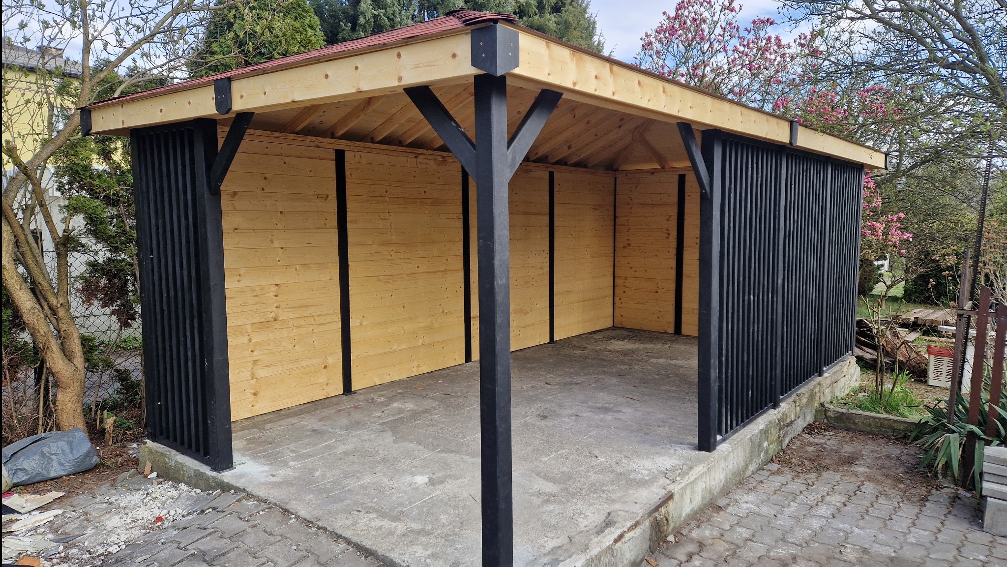 Nowoczesna altana z jasnego drewna i czarnymi elementami konstrukcyjnymi, widok z zewnątrz, betonowa podstawa, drzewa w tle.