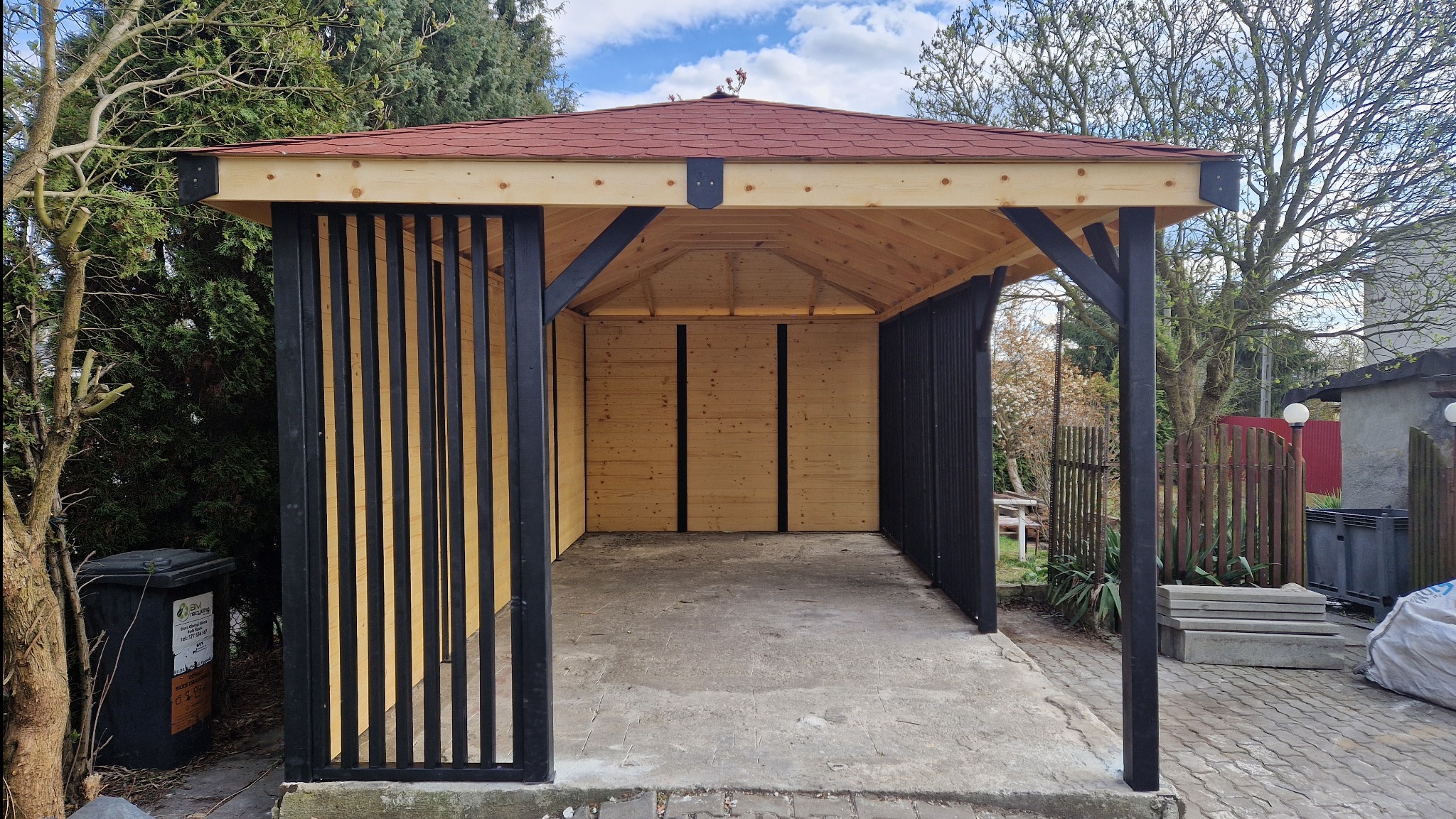 Nowoczesna altana z czerwonym dachem i czarnymi elementami konstrukcyjnymi, otoczona zielenią. Widoczne pionowe lamele i betonowa podłoga.