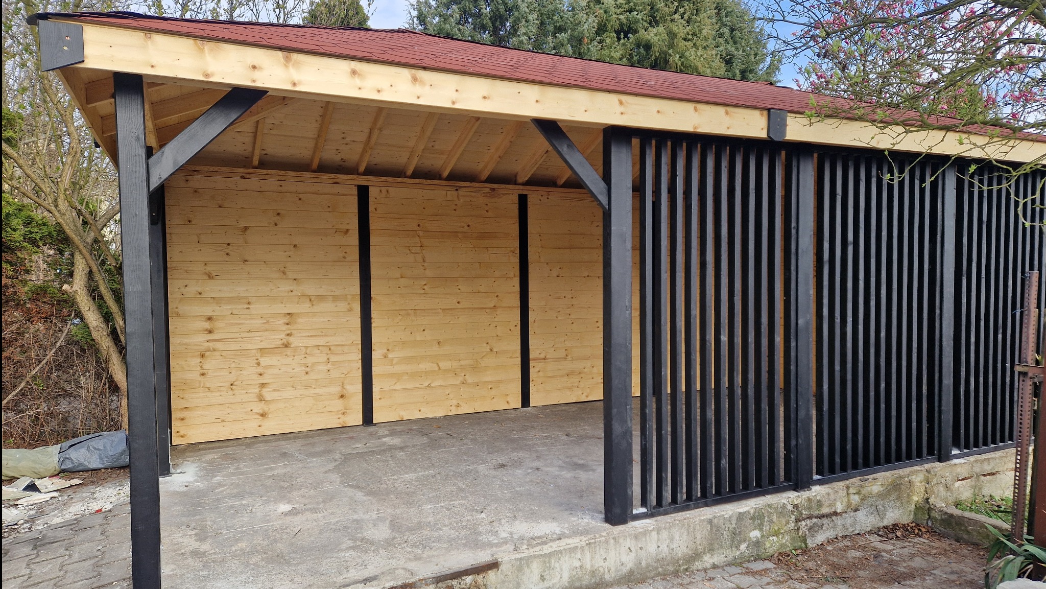 Drewniana wiata z czerwonym dachem, czarnymi słupami i ażurowymi panelami. Konstrukcja z jasnego drewna, betonowa podstawa. Widok na ogród i drzewa.