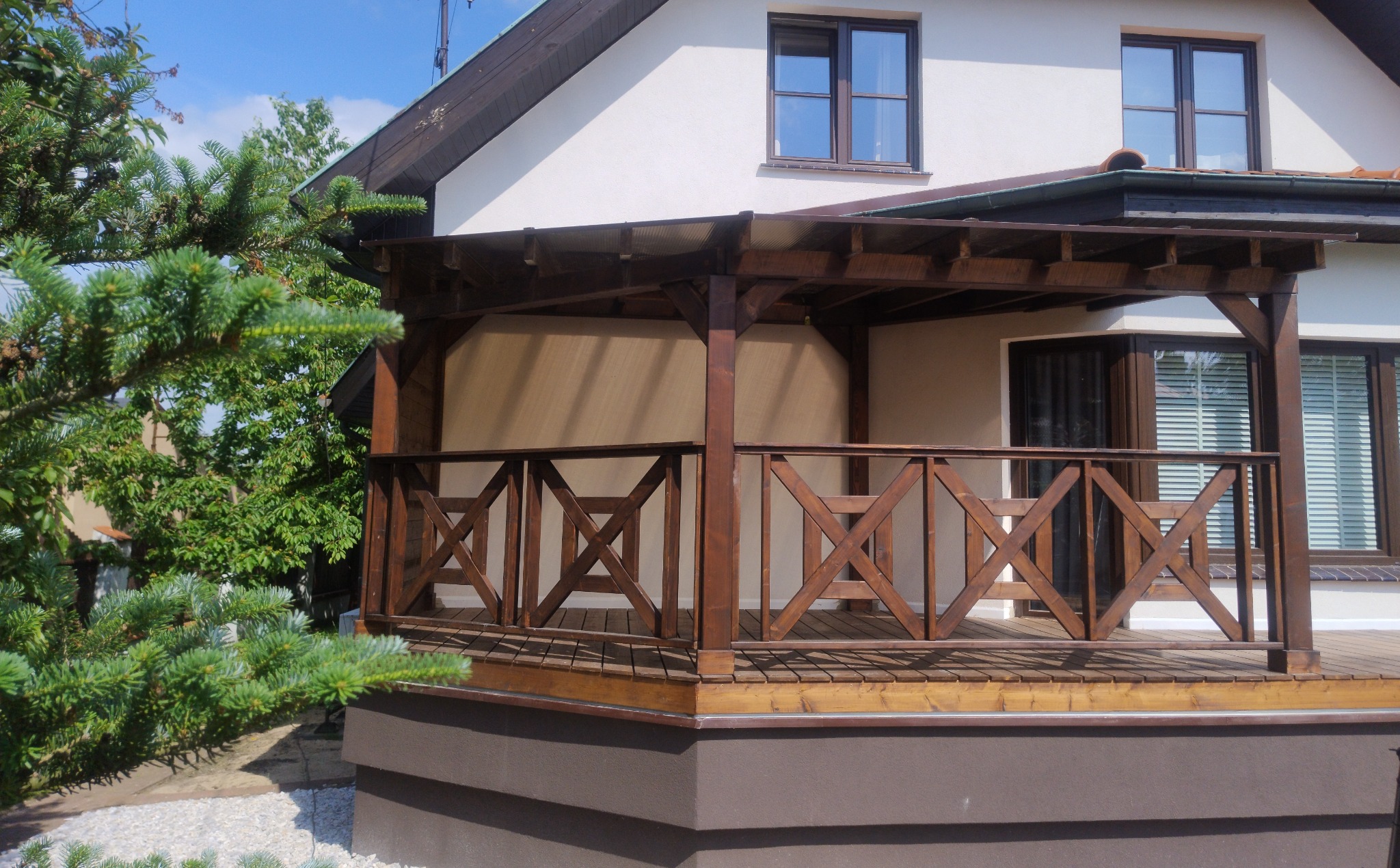 Zadaszony taras z drewnianą balustradą o geometrycznym wzorze X, przylegający do jasnego domu z brązowymi oknami i dachem, obok zielone drzewo iglaste.