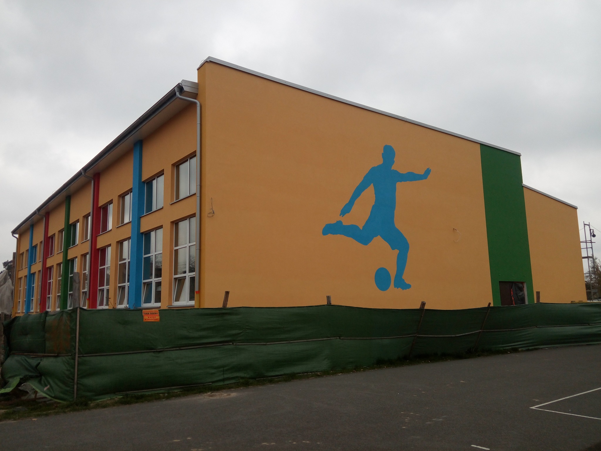 Żółta elewacja szkoły z niebieską sylwetką piłkarza, widoczne rusztowanie i zabezpieczenia terenu budowy.