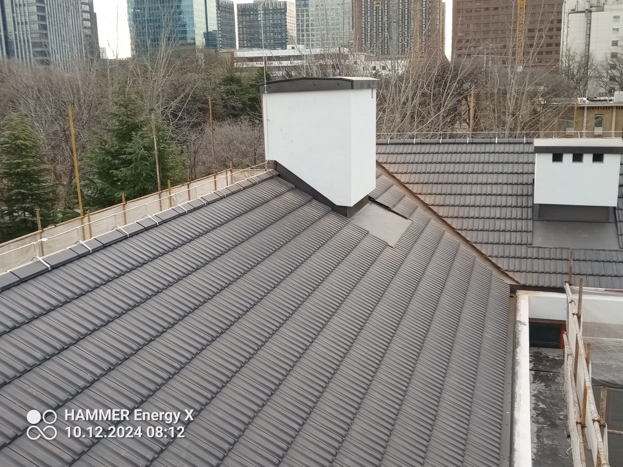 Nowo położony, szary dach z dachówki ceramicznej z białym kominem na tle miejskiego krajobrazu i drzew bez liści.