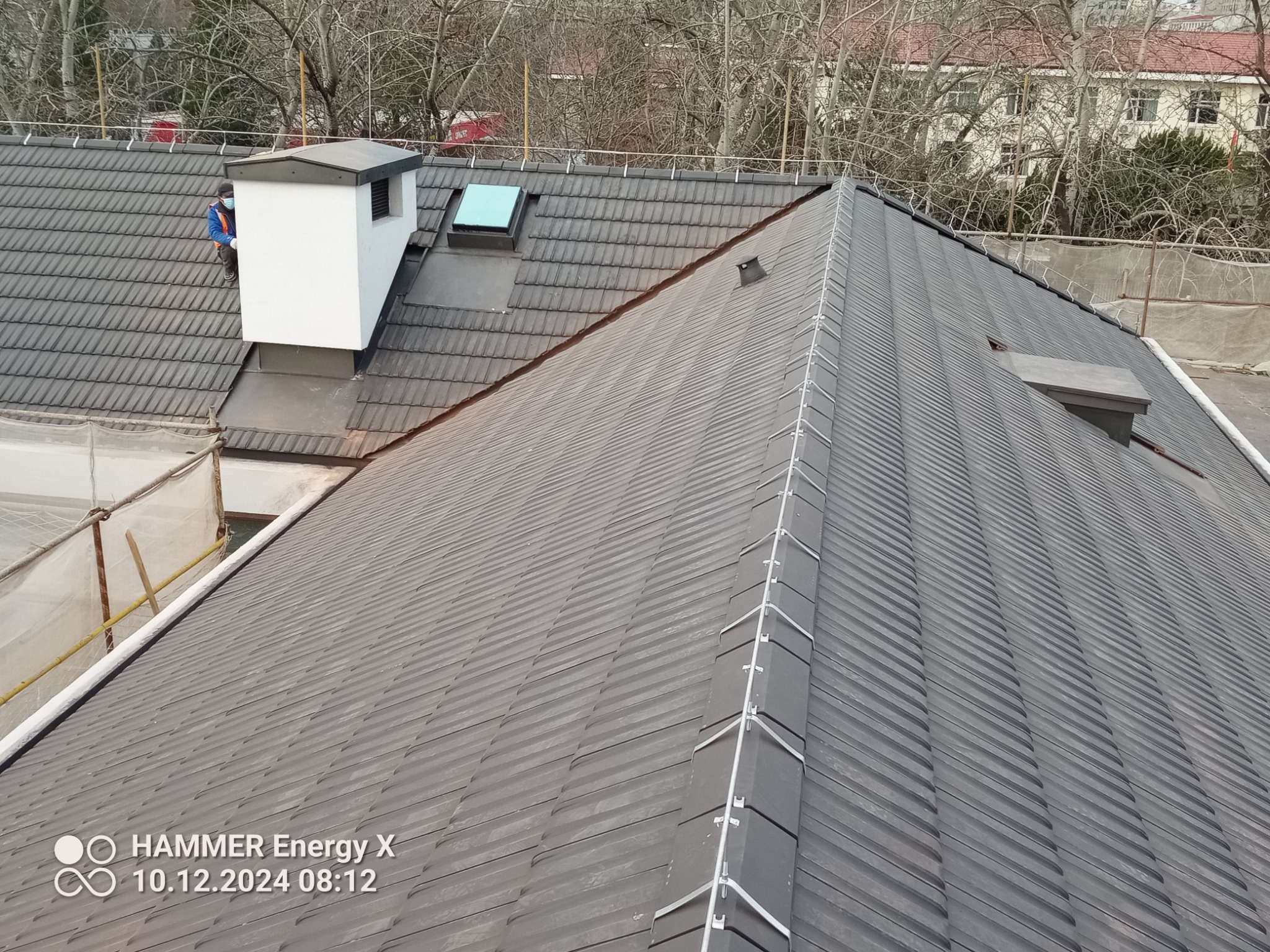 Ukośny widok na szary dach z blachy trapezowej z kominem, świetlikiem i zabezpieczeniami przeciwśniegowymi, w tle drzewa i budynek, widoczny pracownik w masce przy kominie.