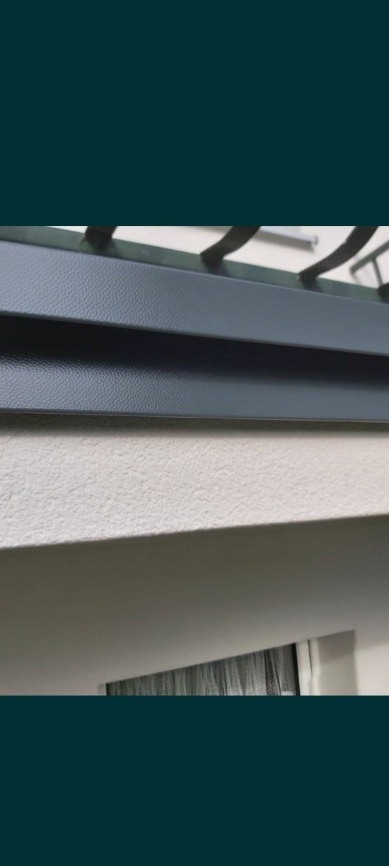 Szczegółowe ujęcie metalowej konstrukcji balkonu z widoczną fakturą i elementami mocującymi, na tle białej ściany i zasłony wewnątrz pomieszczenia.