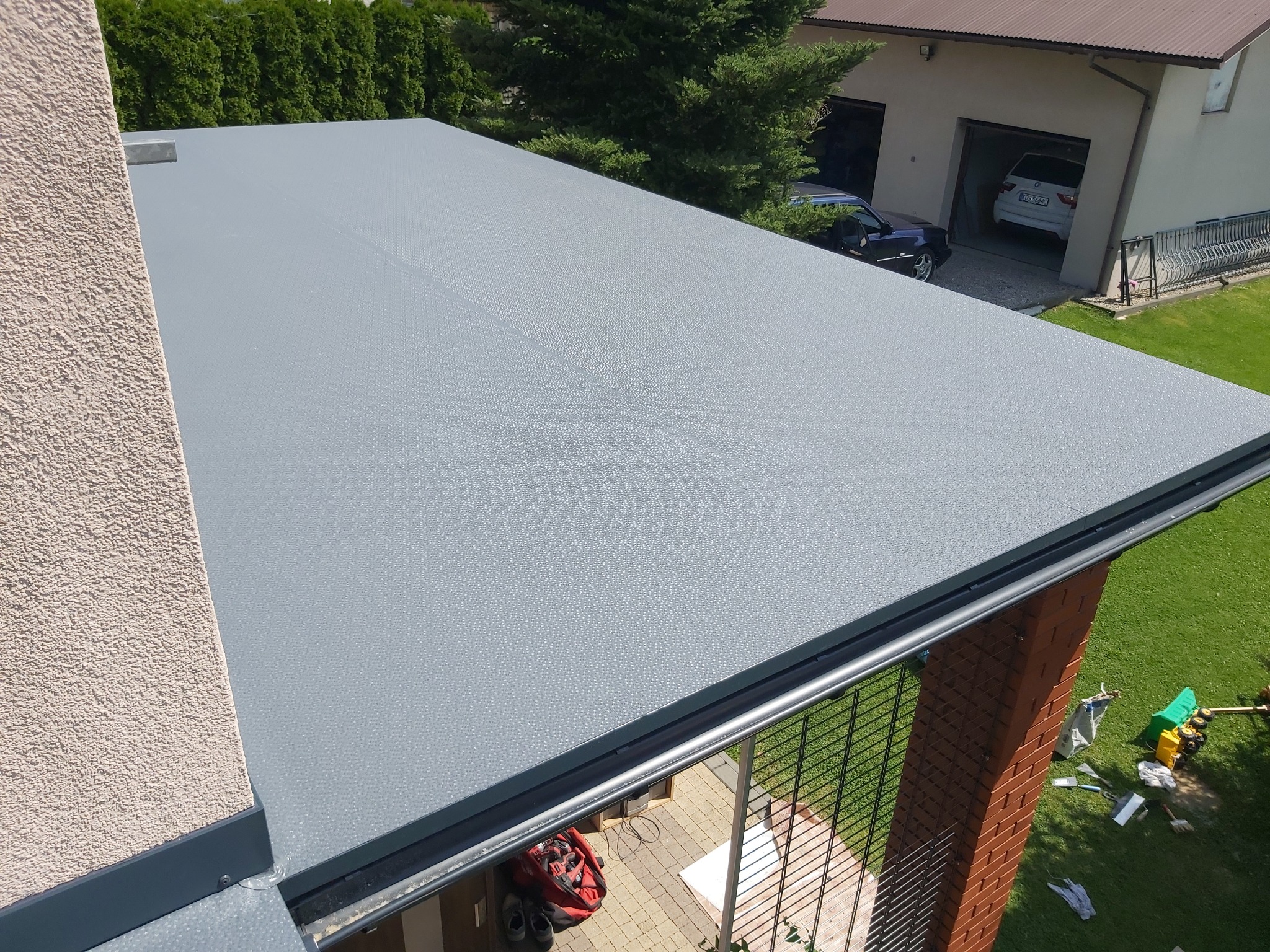 Szary, płaski dach balkonu z widoczną teksturą, z metalowym obramowaniem, sąsiadujący z beżową ścianą budynku. W tle widoczne drzewa, garaż z dwoma samochodami oraz trawnik.