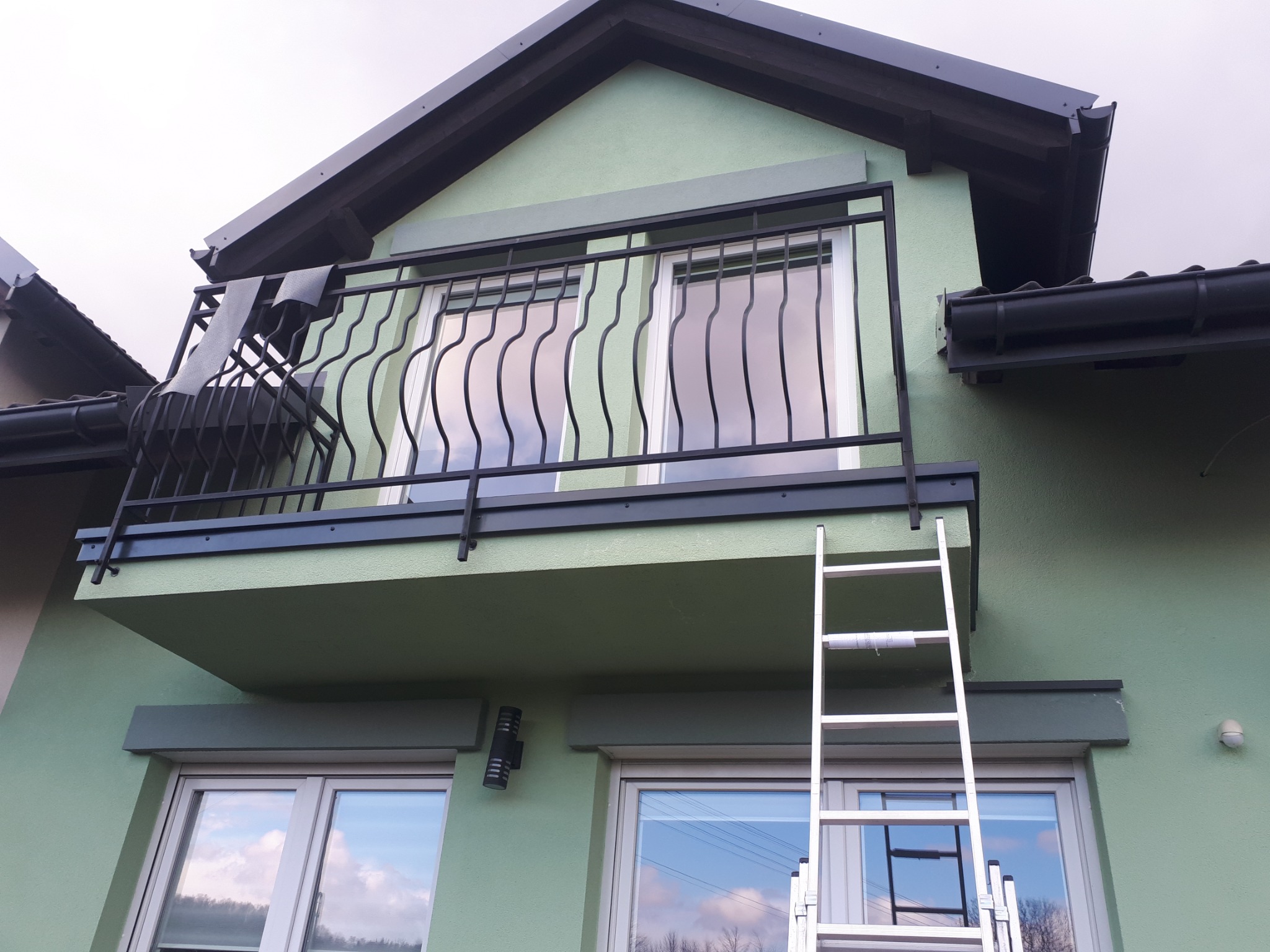Zewnętrzny widok balkonu z czarną, dekoracyjną balustradą, aluminiową drabiną opartą o ścianę i widocznym krzesłem na balkonie, na tle zielonej elewacji budynku.