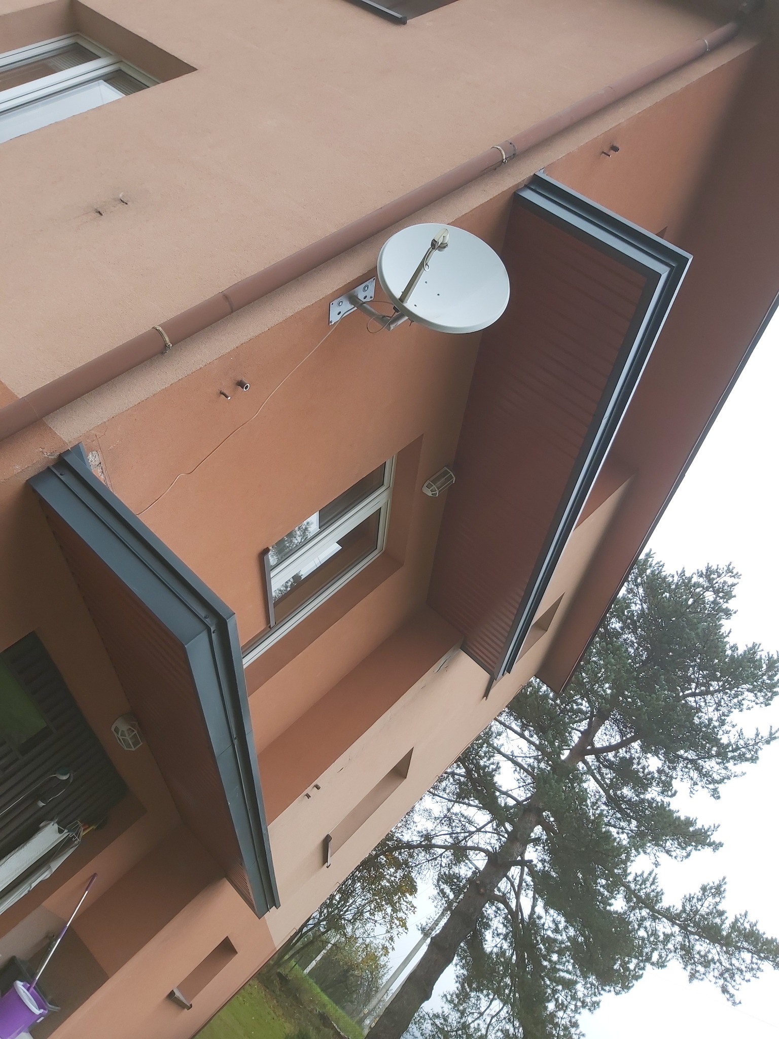 Zewnętrzny widok pomarańczowego budynku z oknami i osłoniętym balkonem z szarą ramą, na ścianie antena satelitarna.