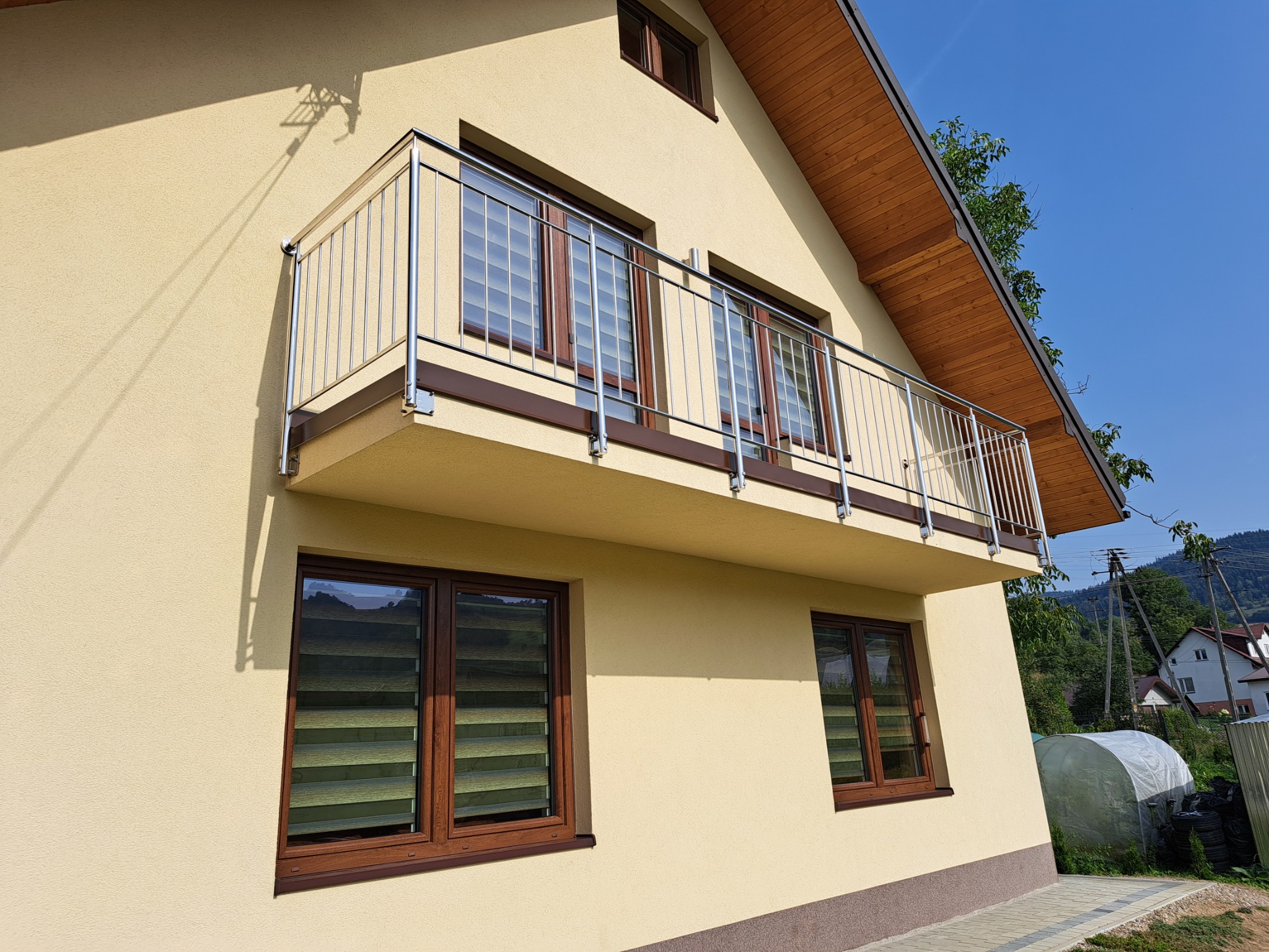 Elewacja domu z balkonem z chromowanymi barierkami, brązowe okna z roletami, widok z dołu na błękitne niebo i zielone drzewa.