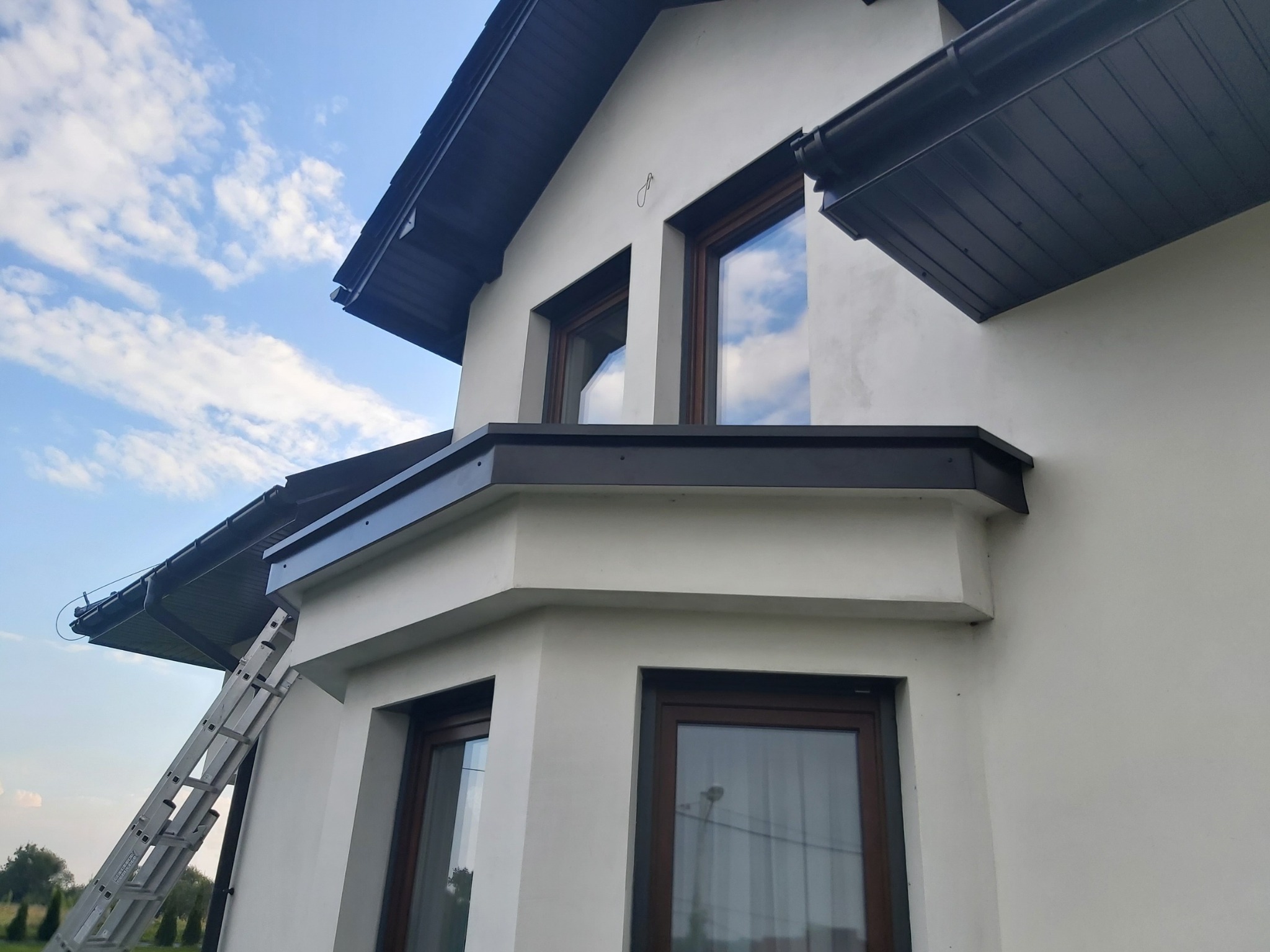 Fragment elewacji domu z oknami w brązowych ramach, widoczna drabina oparta o ścianę, odcień bieli ścian kontrastuje z ciemnym kolorem dachu i rynien.
