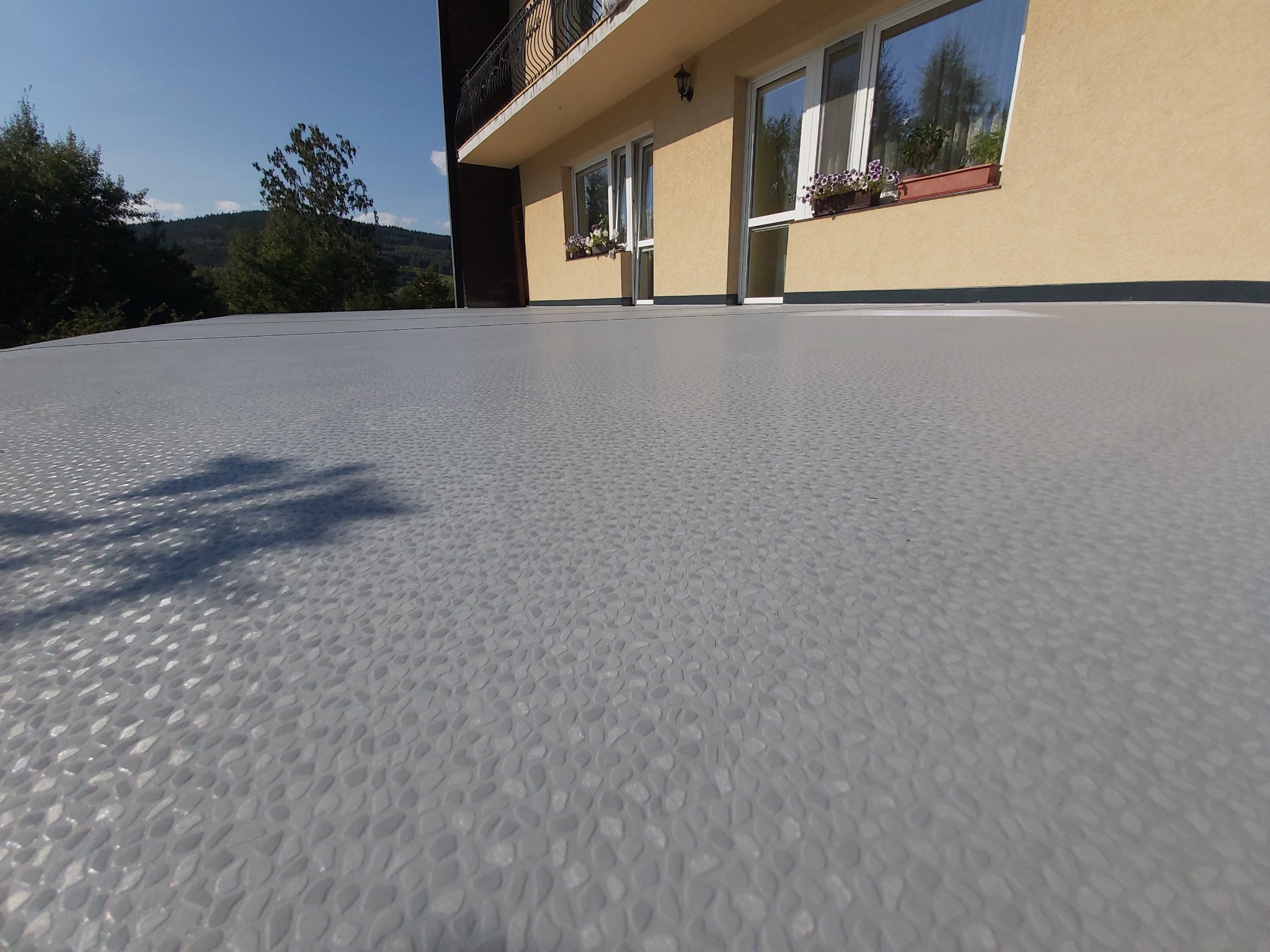 Szary balkon z widoczną fakturą imitującą kamień, z kwiatami w doniczkach na parapecie i widokiem na zalesione wzgórza w tle.