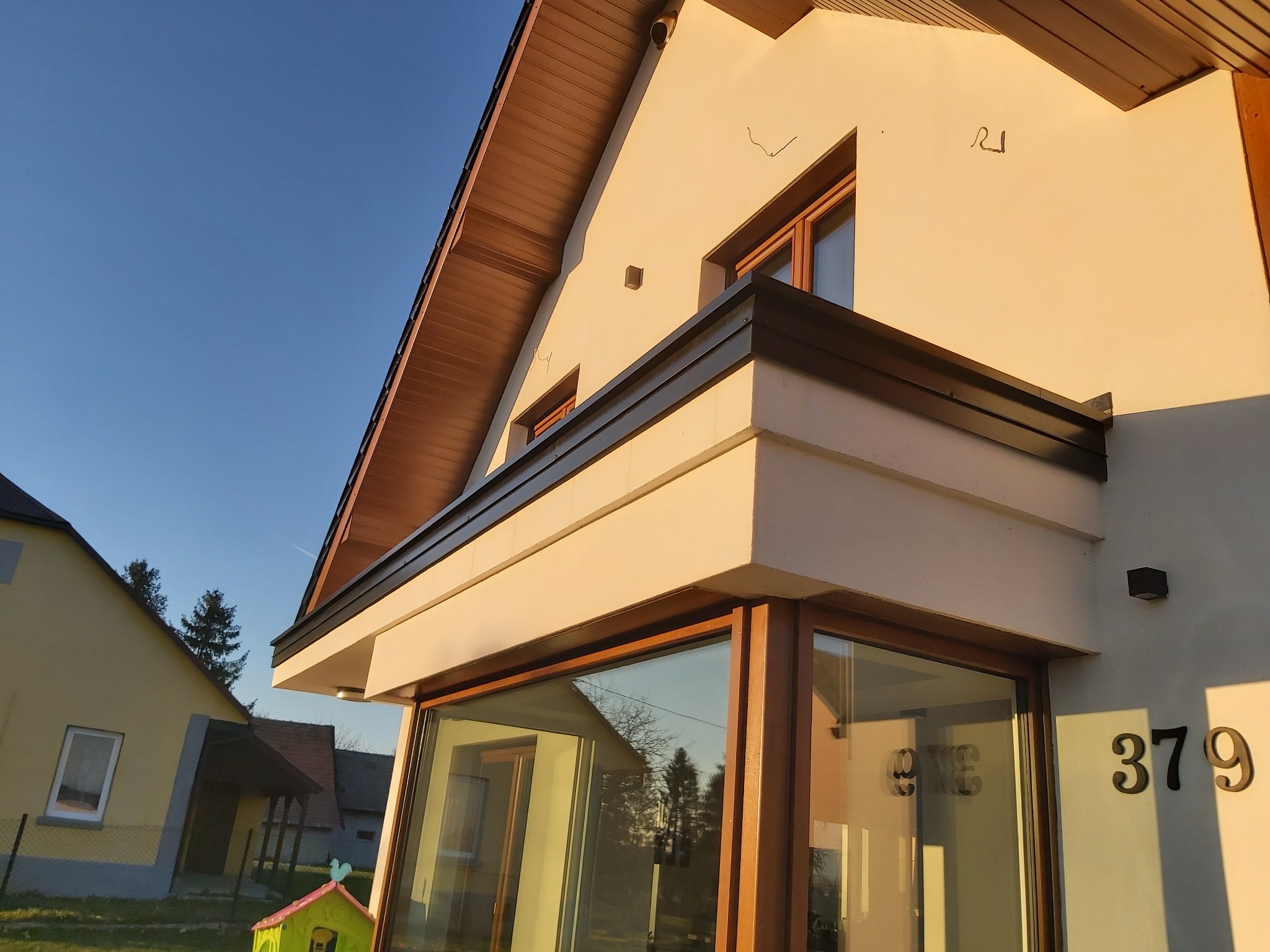 Nowoczesna bryła domu z przeszklonym balkonem, widok z dołu w popołudniowym słońcu, widoczne numery 379 na fasadzie.
