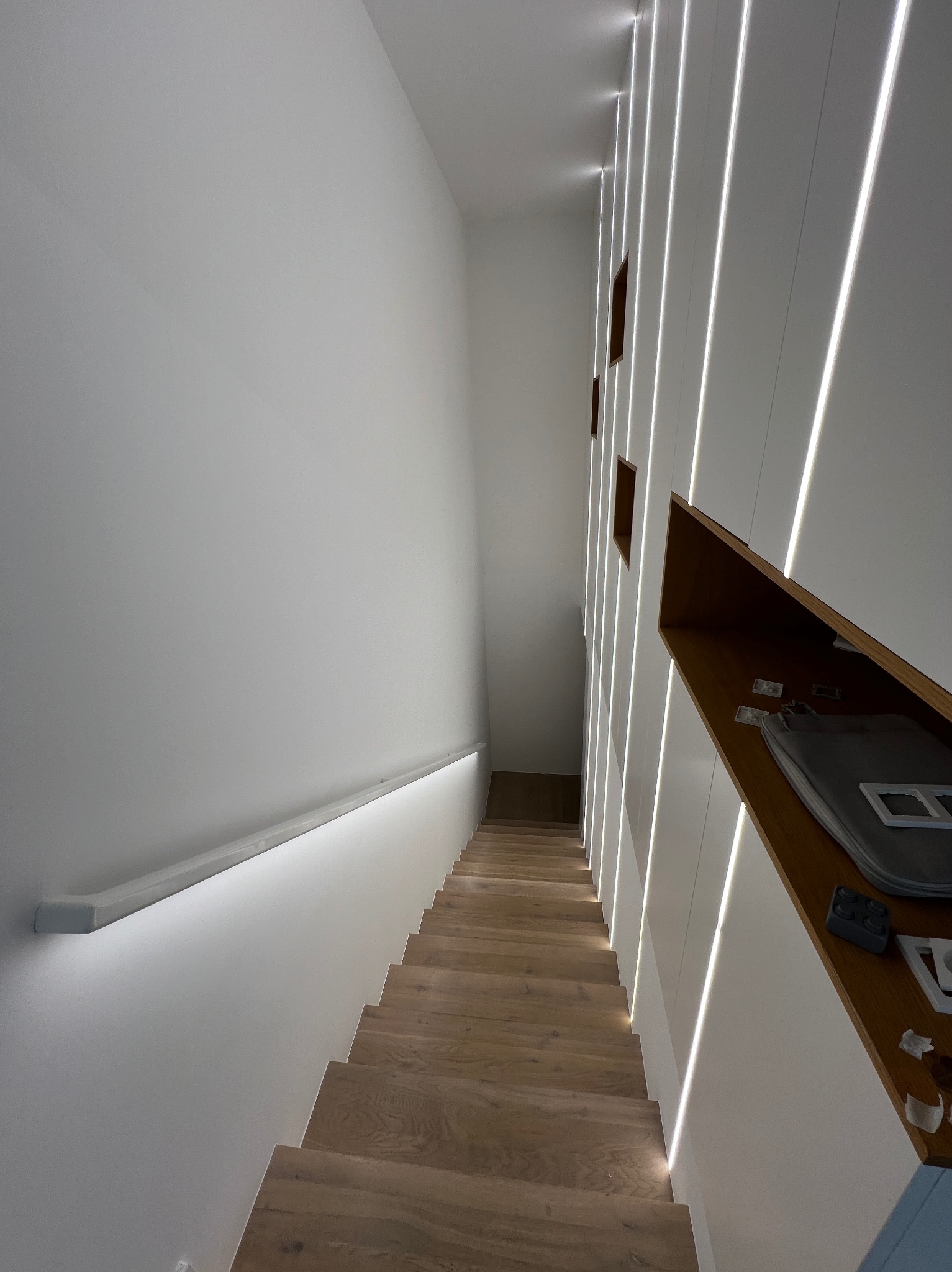 Nowoczesne schody z drewnianymi stopniami, oświetleniem LED wzdłuż ściany i poręczą z podświetleniem, widok z góry w dół, na półce obok schodów leżą narzędzia i akcesoria.