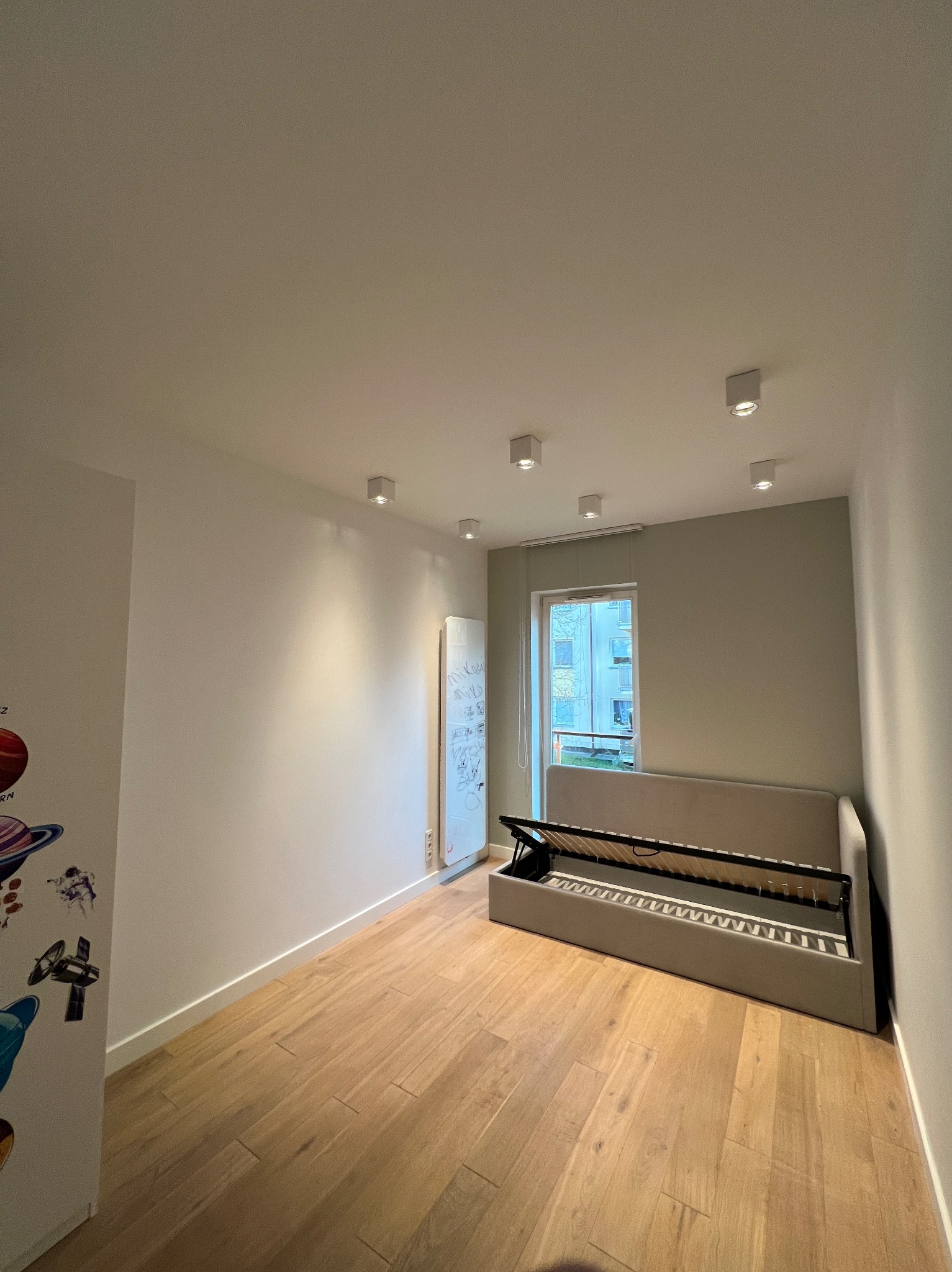 Pokój po remoncie z drewnianą podłogą, otwartym łóżkiem z pojemnikiem i białą tablicą z rysunkami.